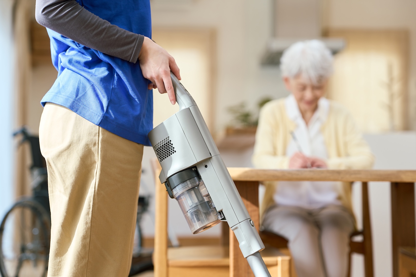 A caregiver vacuums while a senior woman sits at the table.