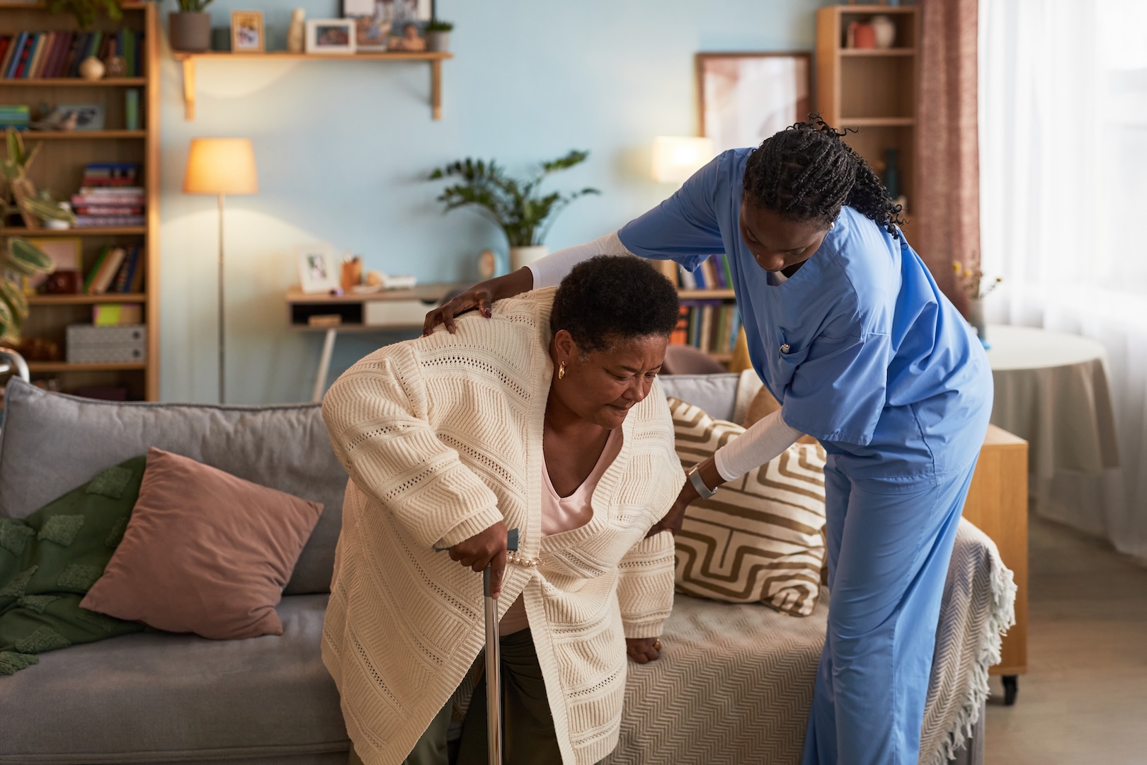 A caregiver helps a senior woman stand from the couch.
